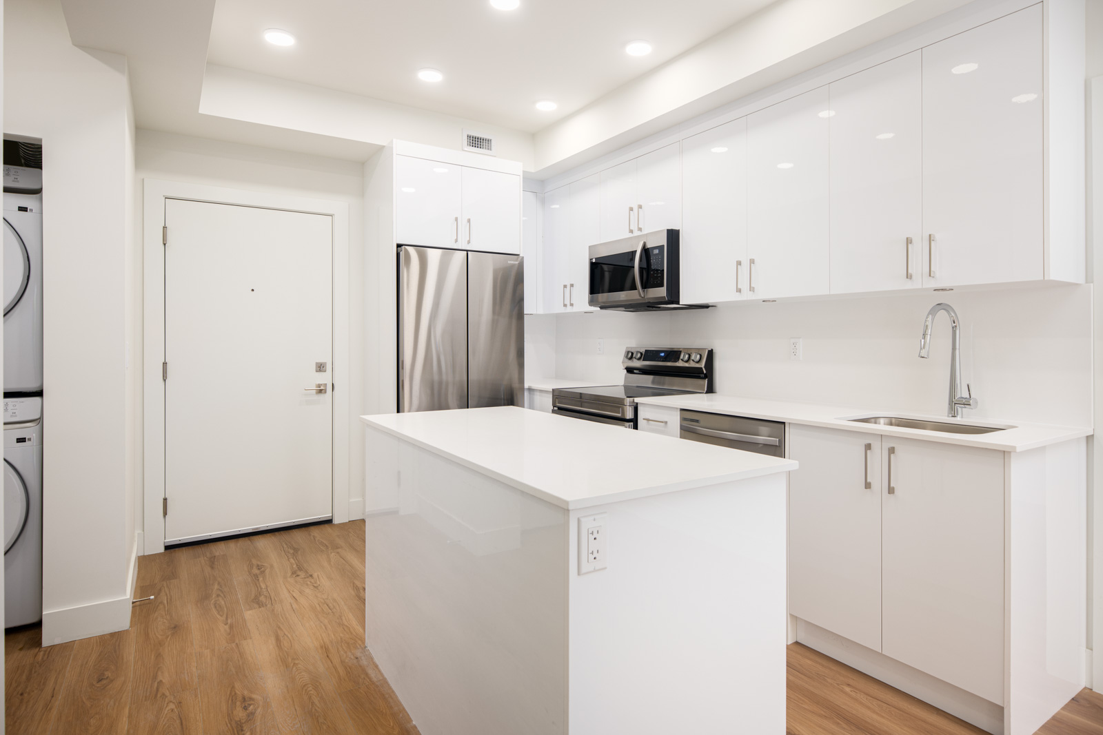 Modern kitchen with white cabinets, stainless steel appliances, an island countertop, wood flooring, and stacked washer and dryer near the entrance.