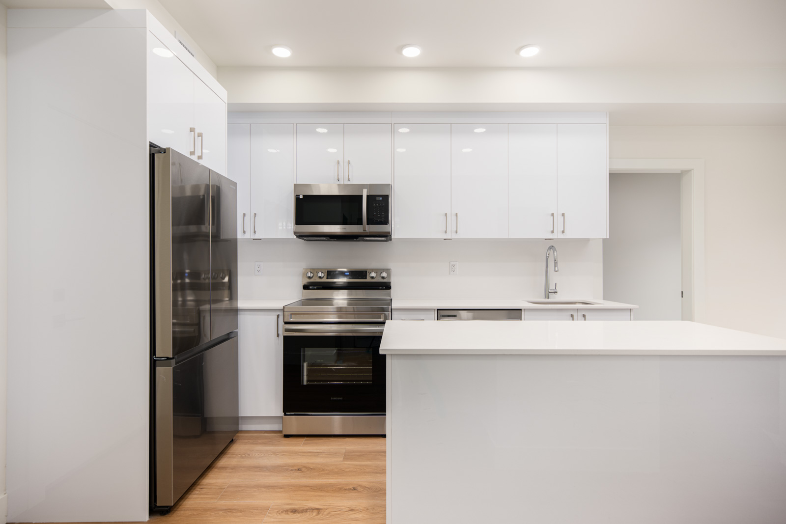 Modern kitchen with white cabinets, stainless steel appliances, electric stove, microwave, and a large white island, set on light wood flooring under recessed ceiling lights.