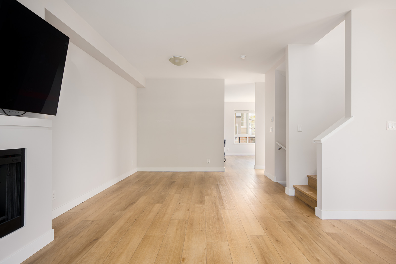Empty living space with light wood flooring, white walls, a fireplace on the left, and stairs on the right leading to an upper floor.