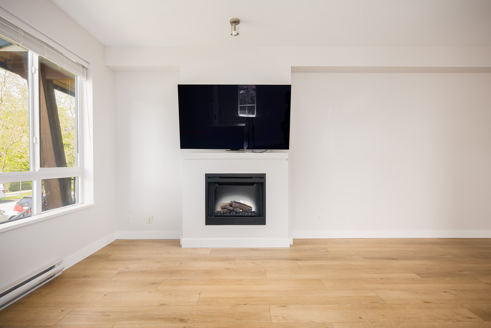 A wall-mounted TV above a modern fireplace in a minimalist living room with light wood flooring and a large window.