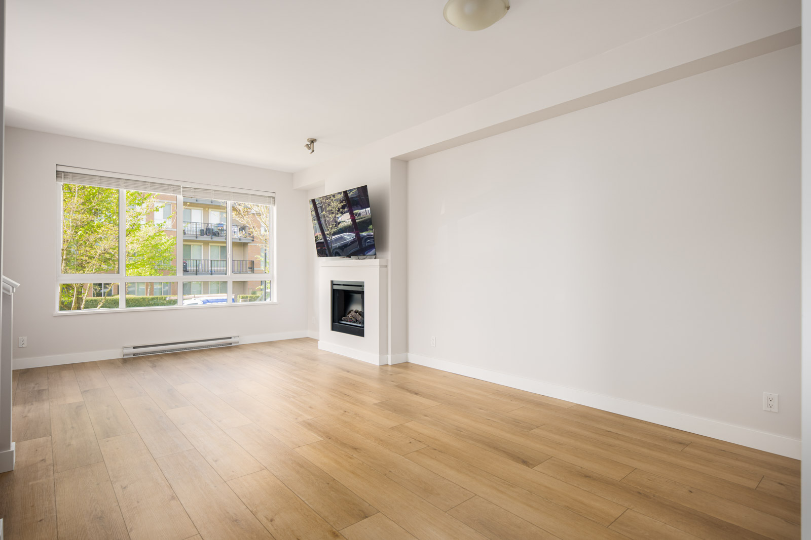 Bright, empty living room with light wooden floors, large window, mounted TV above a fireplace, and white walls.