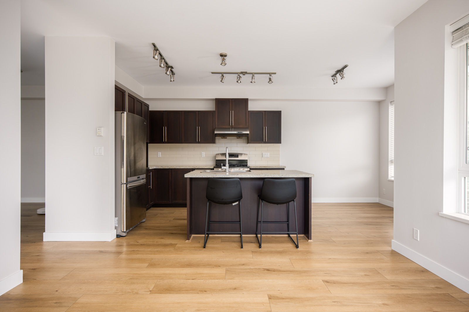 Modern kitchen with dark wood cabinets, stainless steel appliances, a central island with two black chairs, and light wood flooring in a bright, empty room.