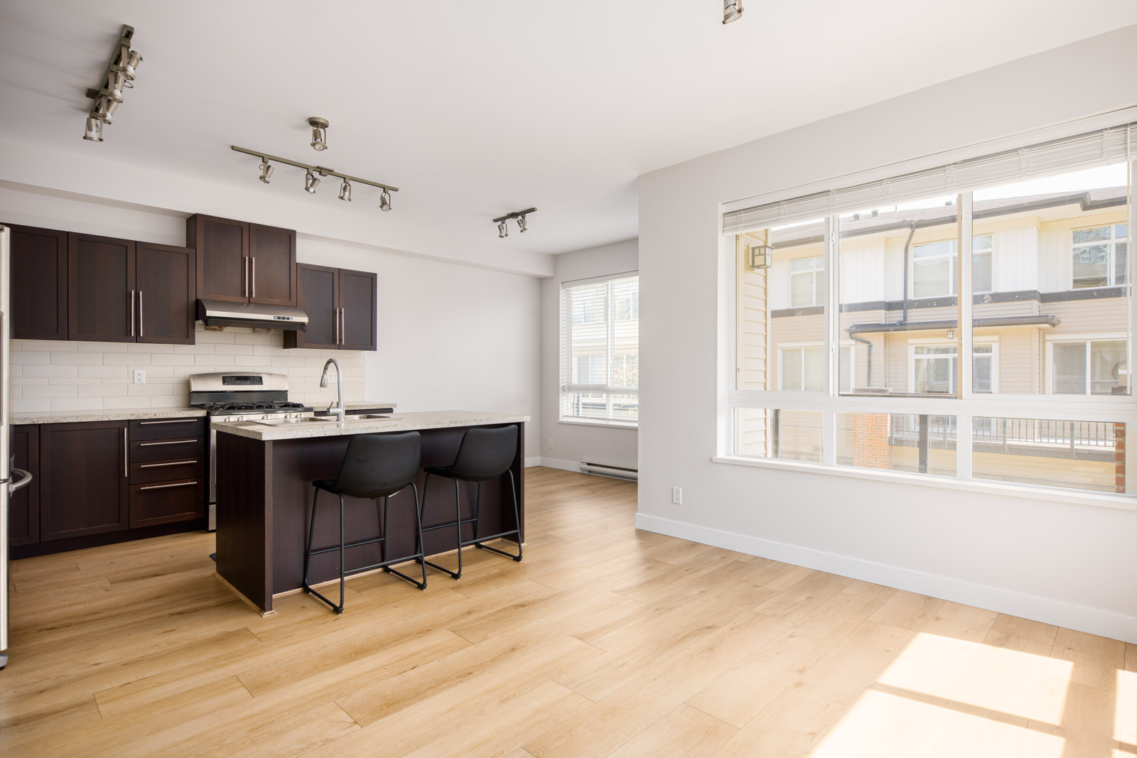 A modern kitchen with dark cabinets, a central island with two black chairs, stainless steel appliances, and large windows letting in natural light.