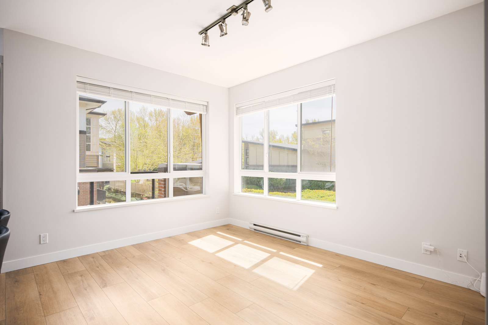 Bright, empty room with light wood flooring, white walls, large windows, and ceiling track lighting, overlooking trees and neighboring buildings.