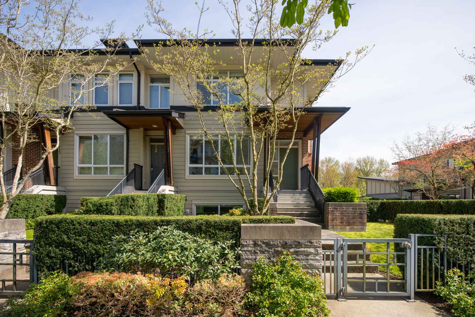 Modern two-story townhouse with large windows, front steps, and landscaped yard with trees and bushes, seen on a sunny day.