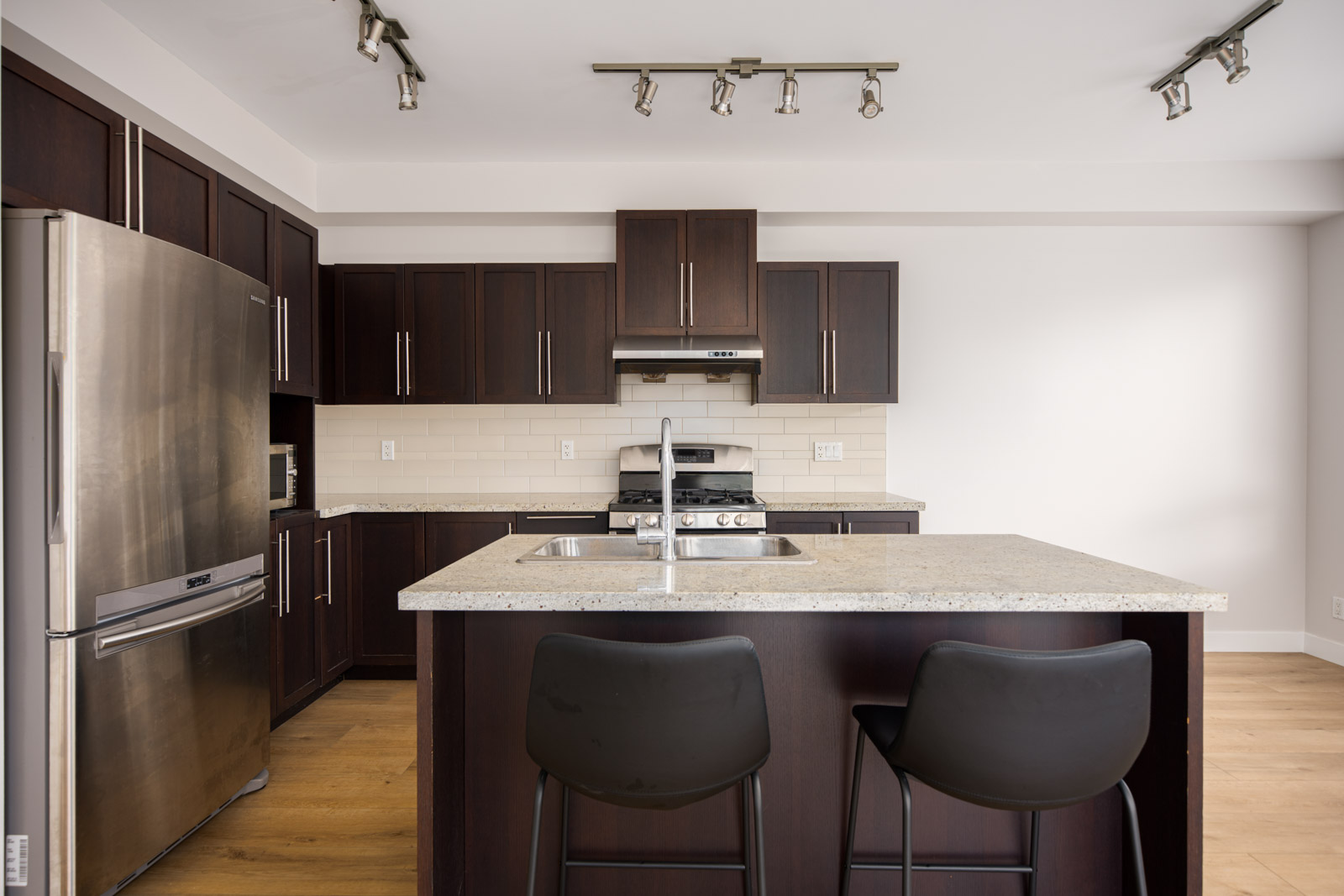 Modern kitchen with dark wood cabinets, stainless steel appliances, a center island with a sink, and two black barstools on a light wood floor.