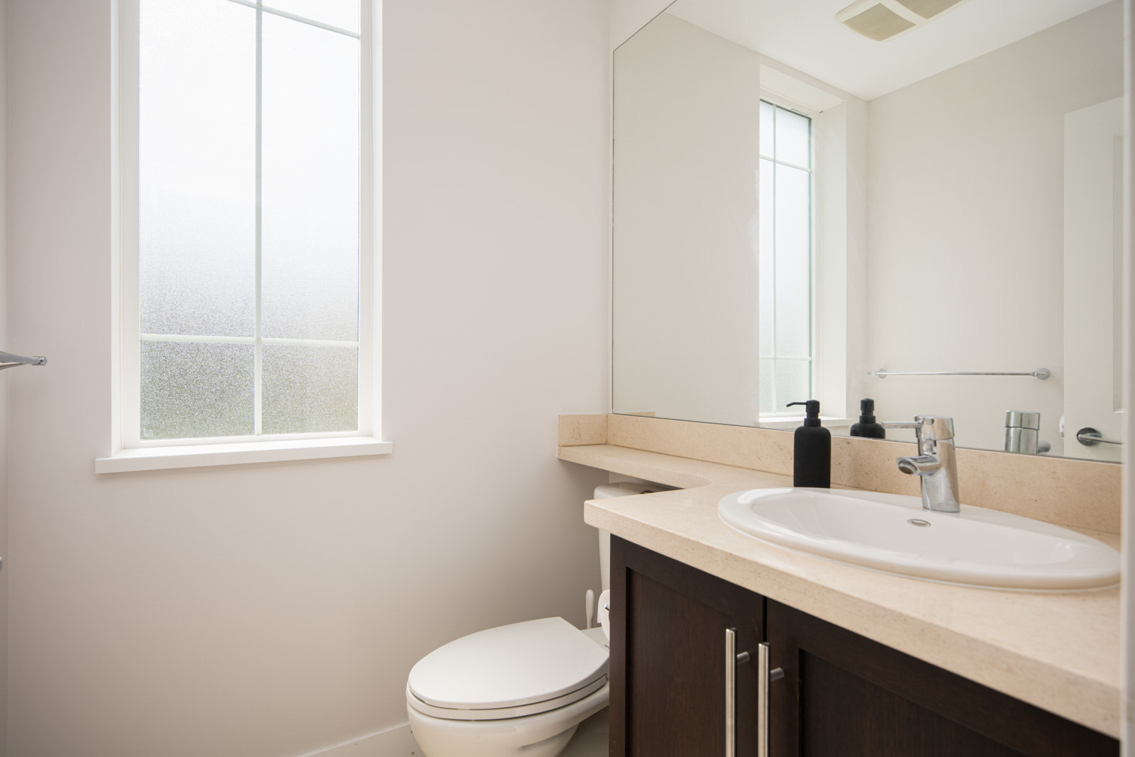 A modern bathroom with a white toilet, a sink on a beige countertop, a large mirror, and frosted window allowing natural light in.