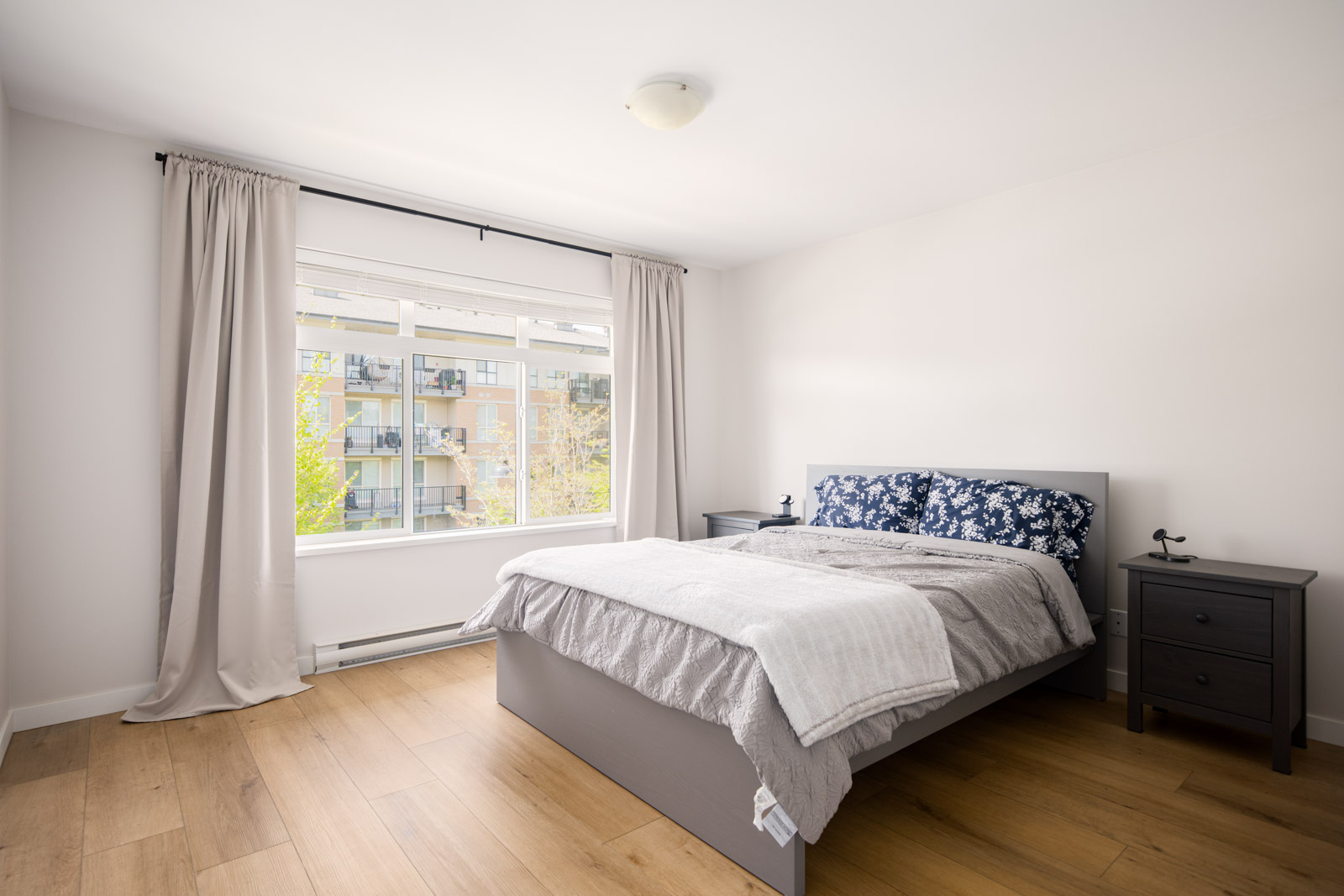 A modern, minimally furnished bedroom with a large window, light wood flooring, a gray bed, matching nightstands, and neutral-colored curtains.