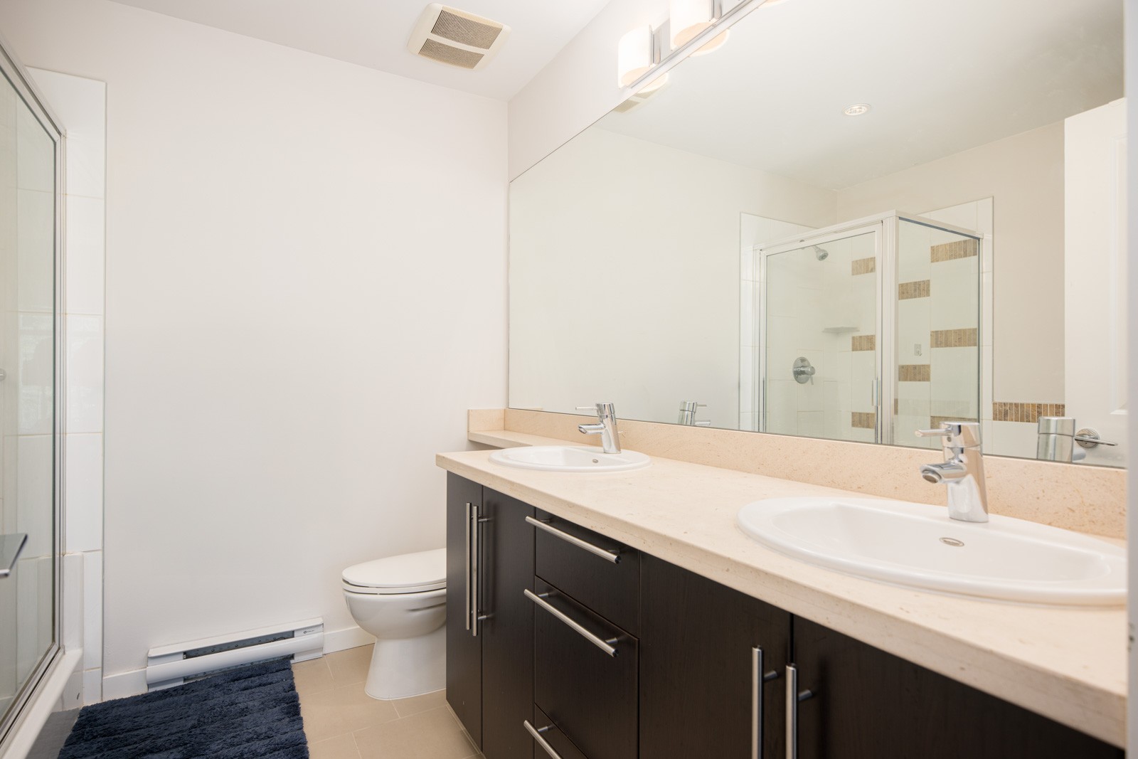 Modern bathroom with a double sink vanity, large wall mirror, toilet, glass-enclosed shower, and a blue rug on a beige tile floor.