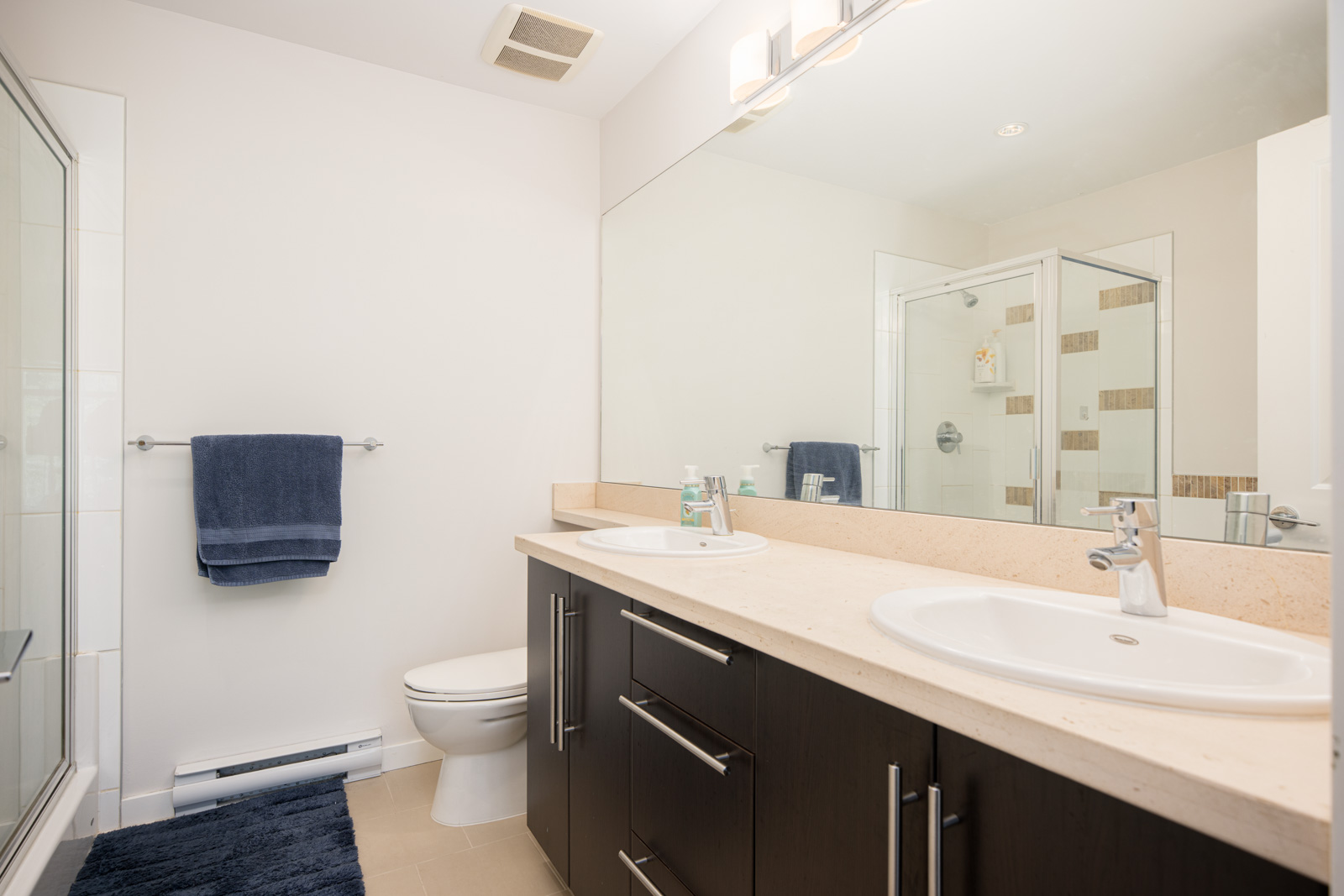 Modern bathroom with double sinks, large mirror, dark wood vanity, toilet, shower with glass door, towel rack with blue towel, and beige floor tiles.