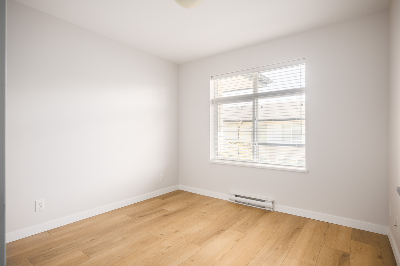 Empty room with light-colored walls, a window with blinds, wooden flooring, baseboard heater, and a ceiling light fixture. Natural light enters through the window.