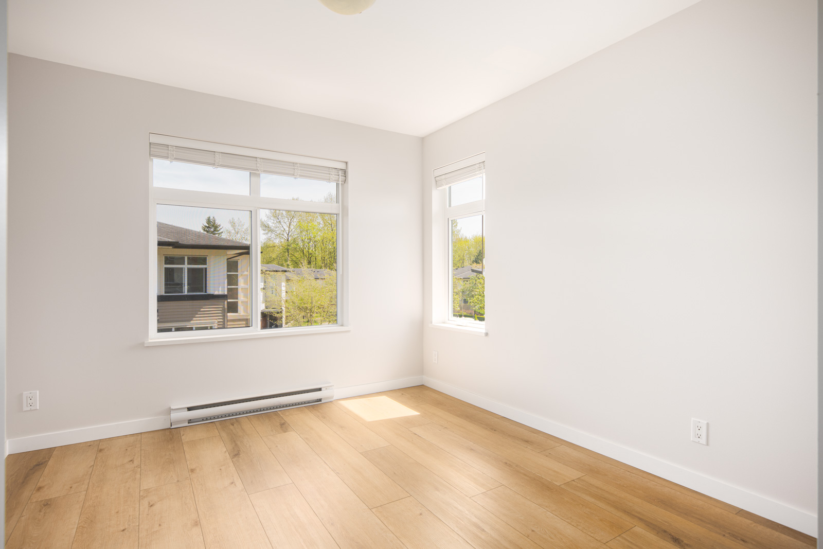 Empty room with light-colored walls, large windows, light wood flooring, and natural light streaming in.