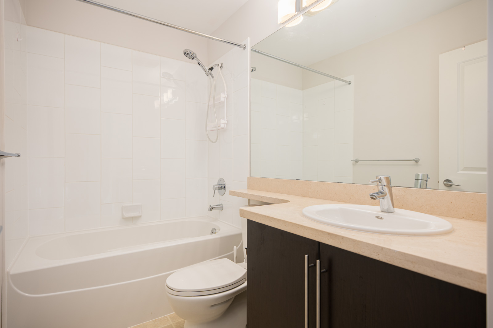 A modern bathroom with a bathtub-shower combo, white sink on a beige countertop, large mirror, and dark cabinets.