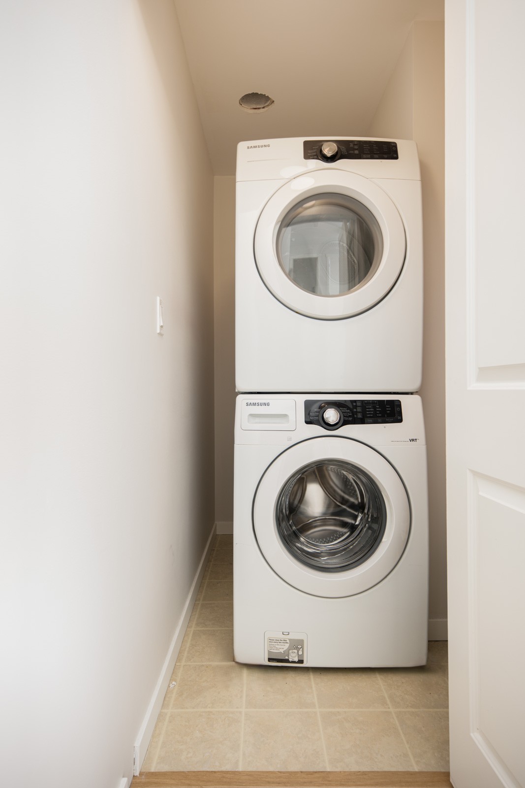 A stacked washer and dryer set in a small laundry nook with beige tile flooring and white walls.