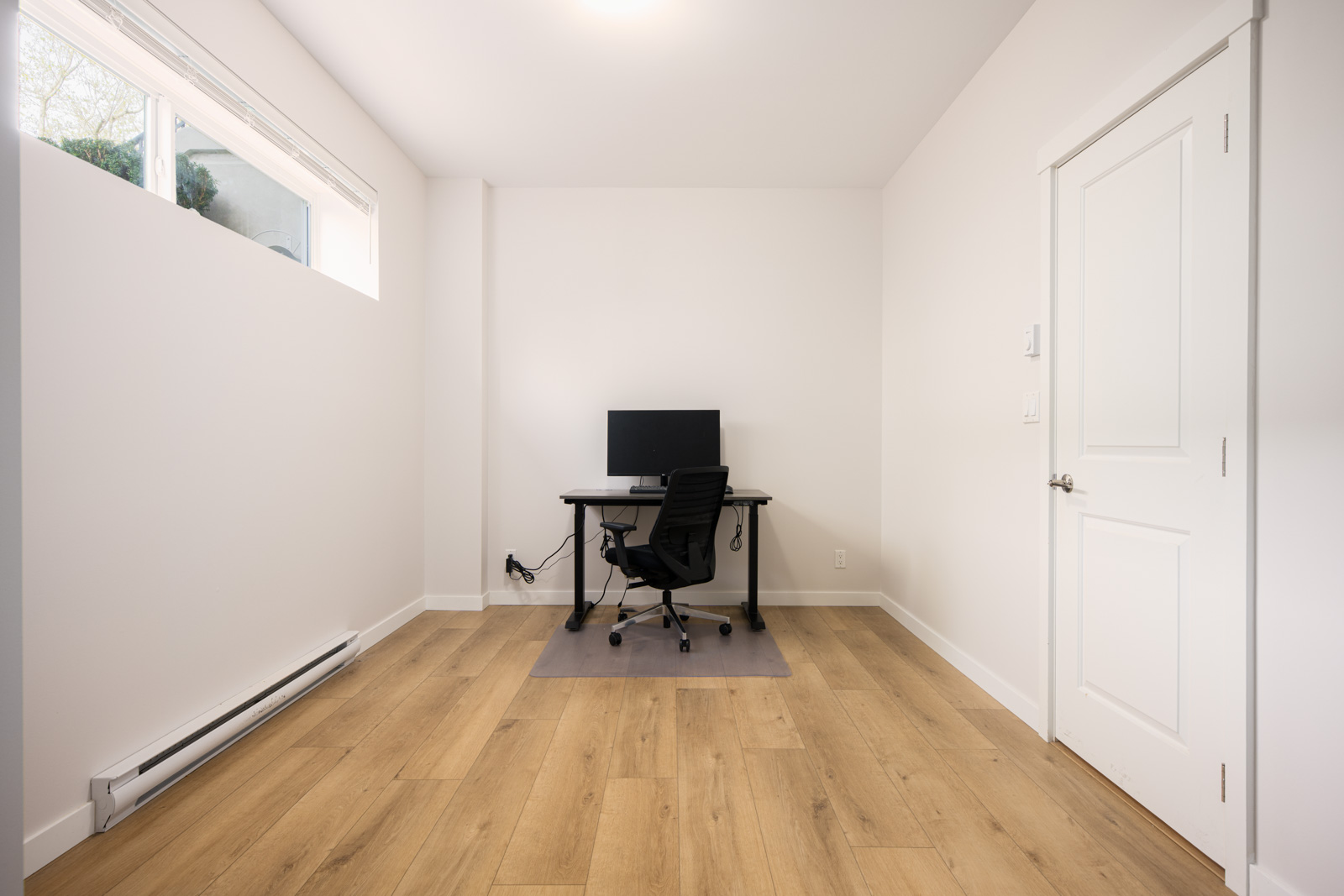 A minimalist office with light wood flooring, a desk with a computer and chair, white walls, and a closed door. Natural light comes from a high window.