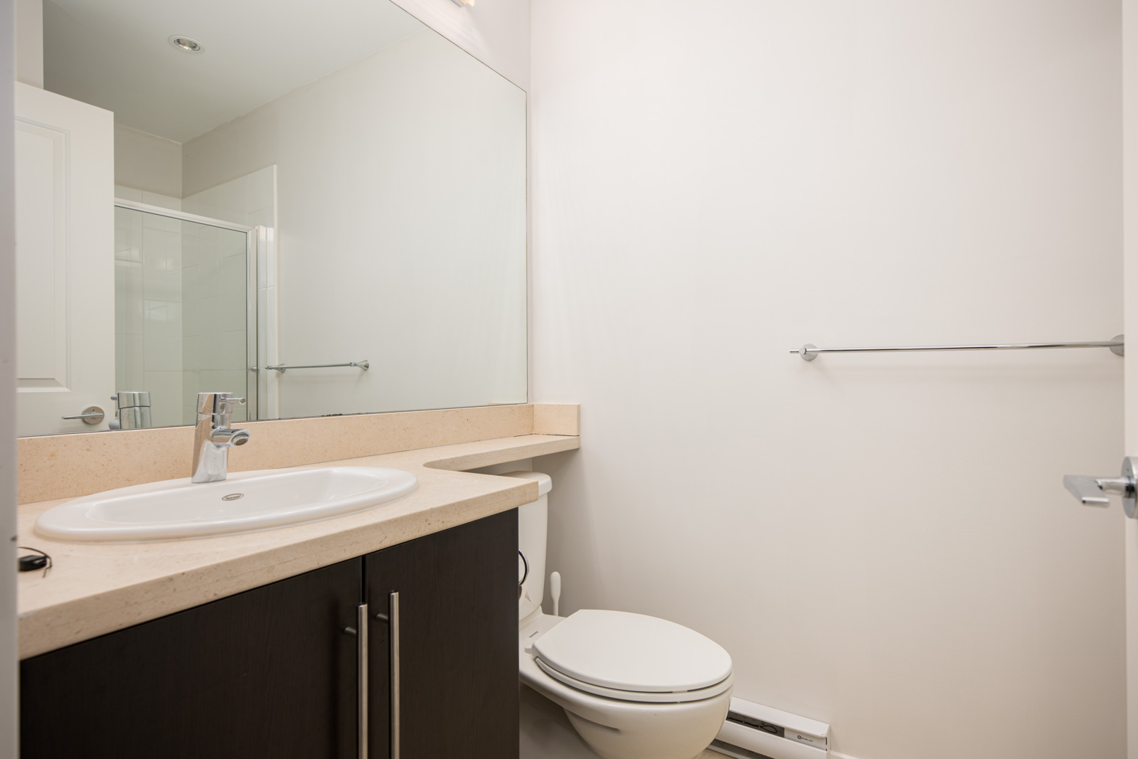 Modern bathroom with a large mirror, white sink on a beige countertop, dark vanity, toilet, towel bar, and glass shower enclosure in the background.