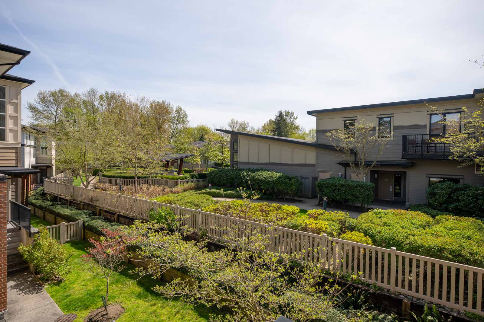 View of a modern residential complex with neatly trimmed bushes, trees, a wooden fence, and a pathway on a sunny day.