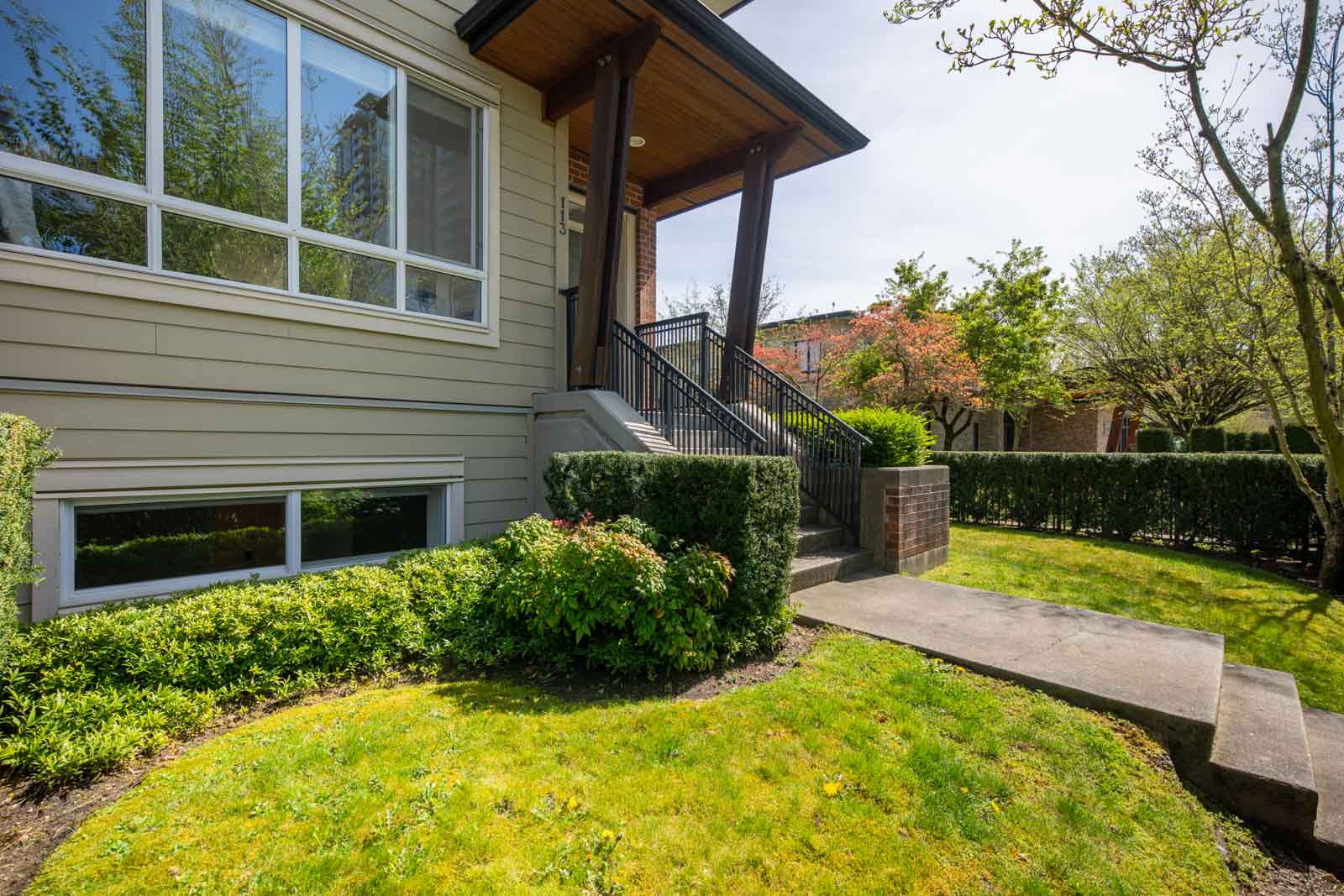 Front entrance of a house with steps leading to a covered porch, large window, manicured lawn, hedges, and trees on a sunny day.