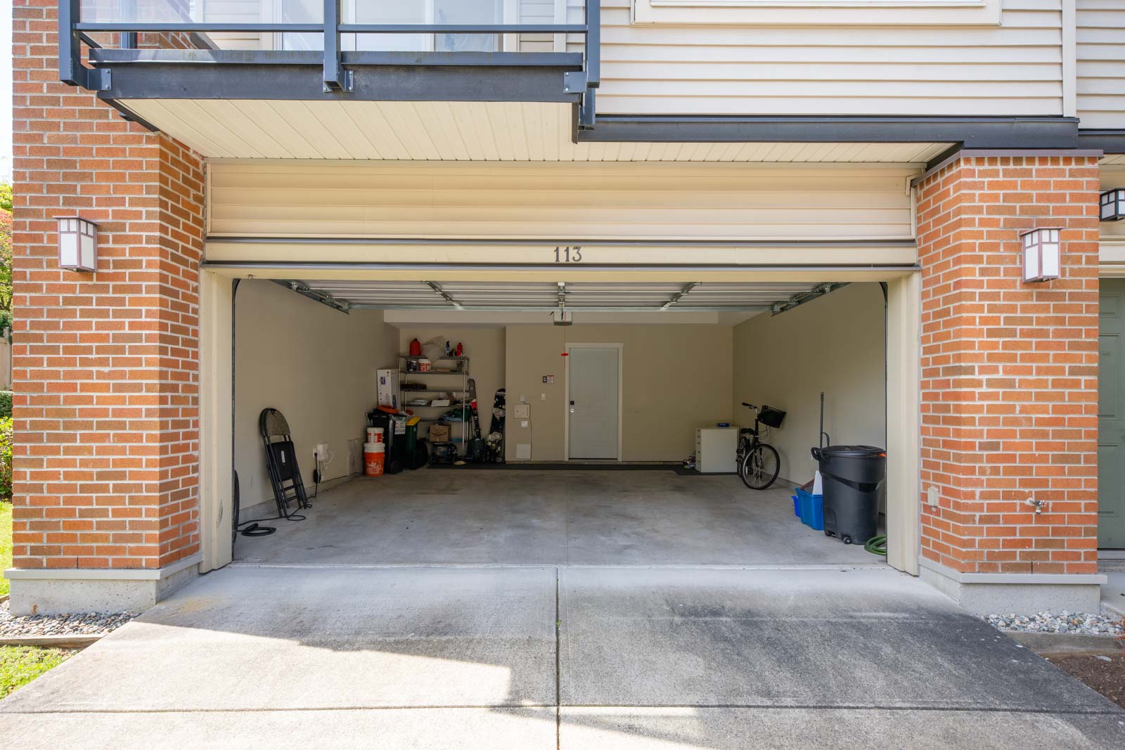 Open two-car garage with beige walls, some shelves, tools, a bicycle, and miscellaneous items stored along the back wall. The garage door is fully open.
