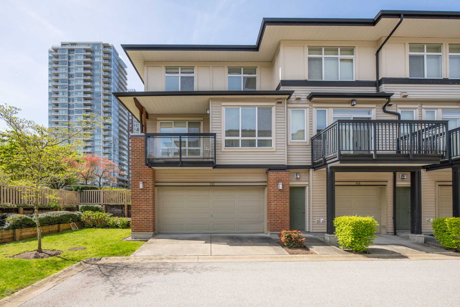 Modern three-story townhouse with a double garage, balcony, and landscaped front yard, adjacent to a high-rise building under a clear blue sky.