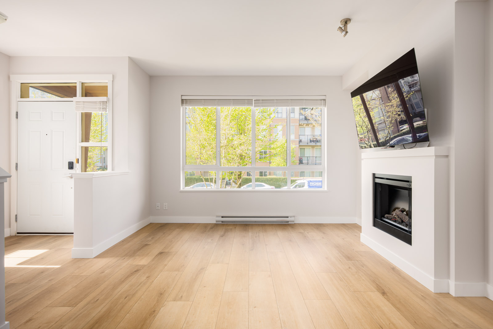 Bright, empty living room with light wood floors, large window, wall-mounted TV above a fireplace, and a white front door.