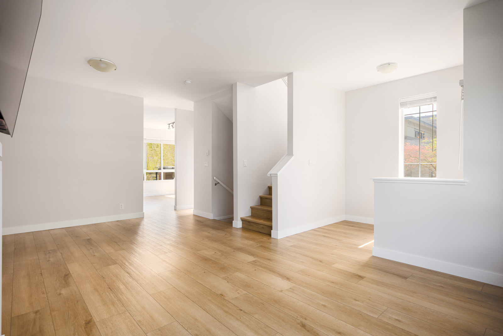 Bright, empty room with light wood floors, white walls, a staircase, and a window letting in natural light. Open layout leads to another room in the background.