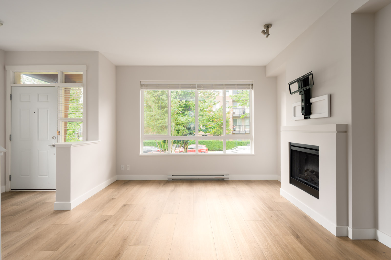Bright, empty living room with light wood flooring, white walls, a large window, fireplace, wall-mounted TV bracket, and a door leading outside.