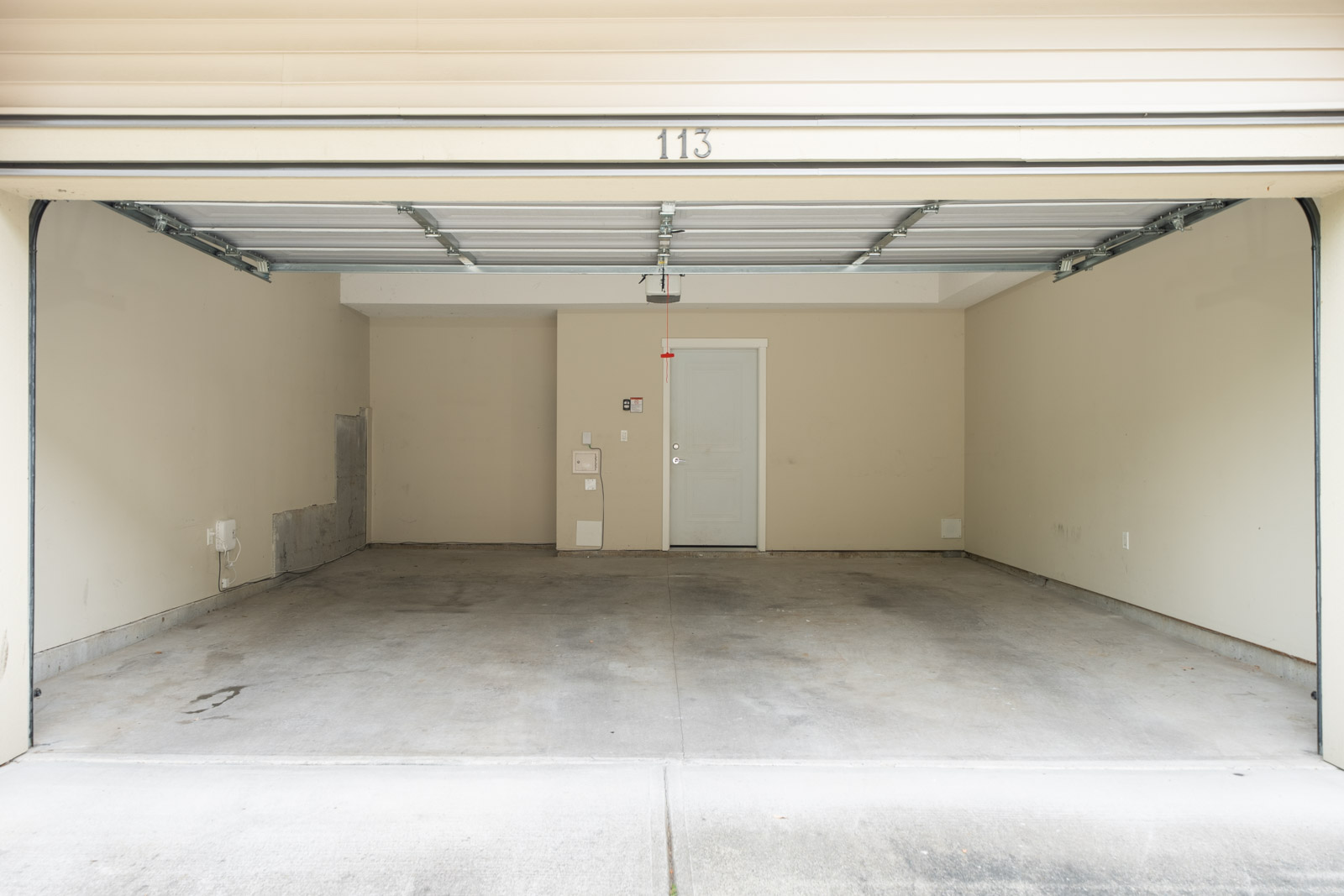 Empty two-car garage with beige walls, a concrete floor, and an open garage door. A single white door is centered at the back wall. The number 113 is displayed above the opening.