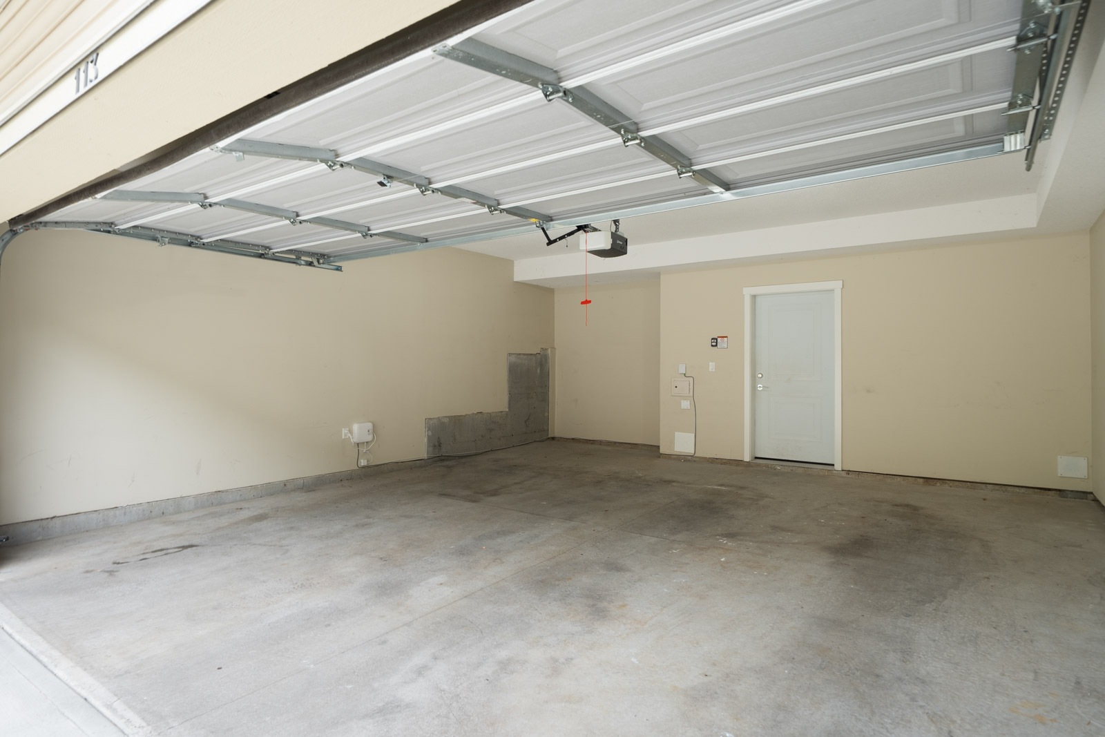 Empty two-car garage with an open door, beige walls, concrete floor, and a white door at the back leading into the house.