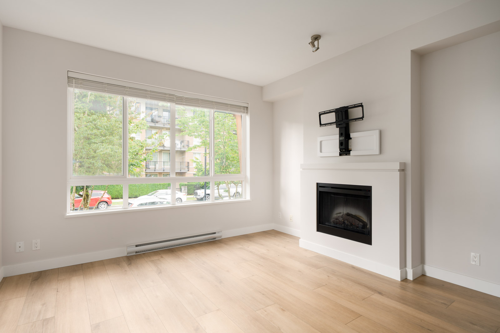 Bright, empty living room with light wood flooring, a large window, and a modern fireplace with a TV mount above it. Trees and parked cars are visible outside.