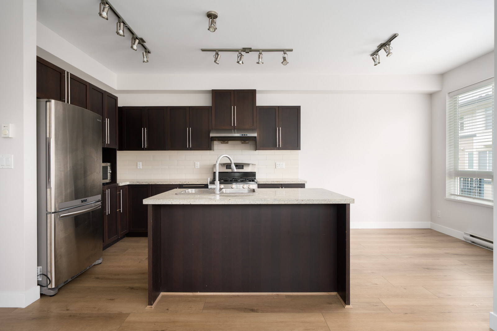 Modern kitchen with dark wood cabinets, stainless steel appliances, a central island with a sink, light wood floors, and large windows letting in natural light.