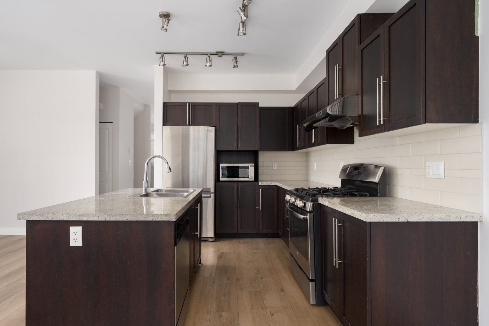Modern kitchen with dark wood cabinets, stainless steel appliances, a central island with sink, and light-colored countertops and backsplash.