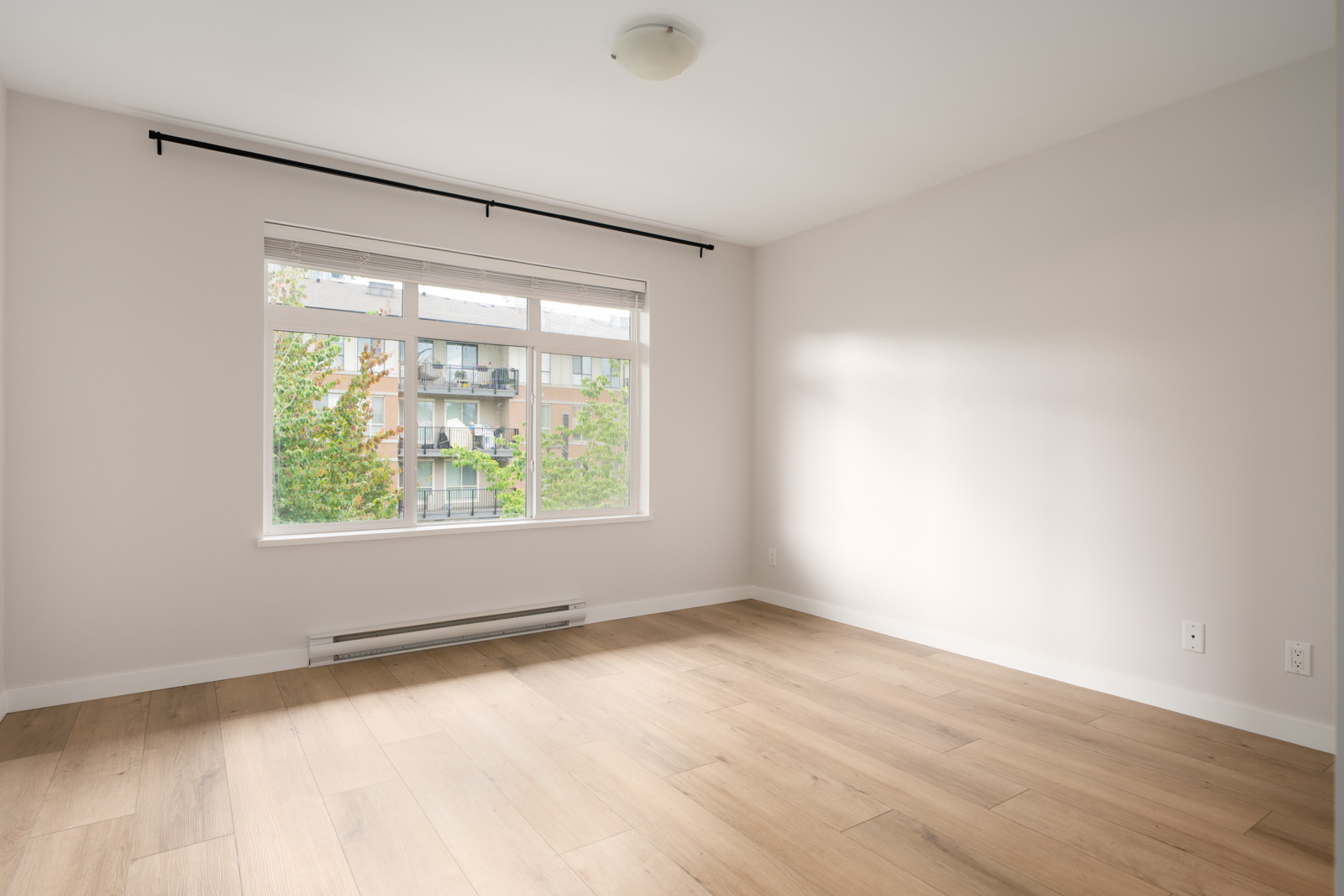 Empty room with light-colored walls, large window with a view of an apartment building, wooden floor, and ceiling light fixture.