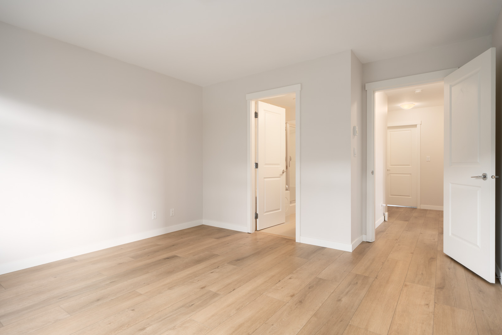 Empty room with light wood flooring, white walls, two open doors leading to a bathroom and a hallway, and natural light coming in from the left.