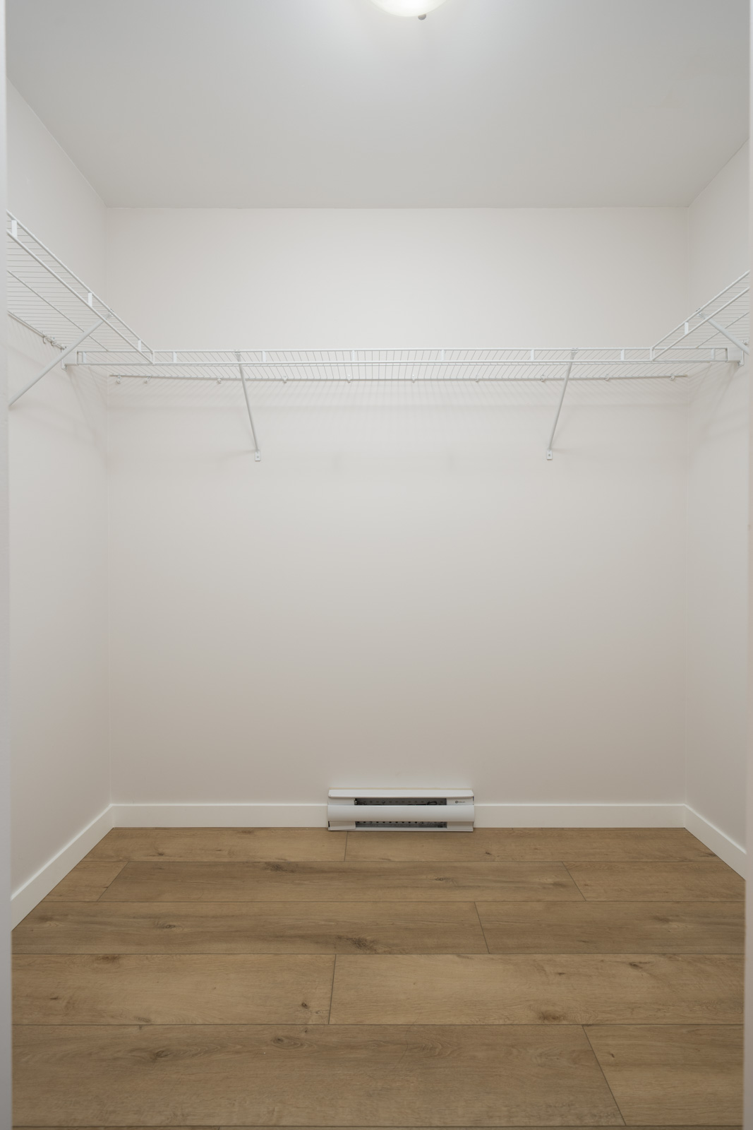 Empty walk-in closet with light-colored walls, wooden floor, overhead wire shelving, and a baseboard heater on the back wall.