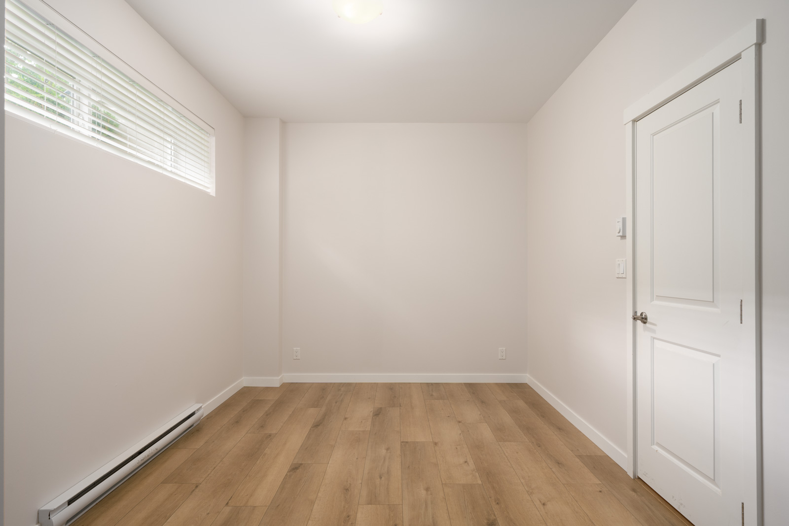 Empty room with light-colored walls, a closed white door, wood flooring, a baseboard heater, and a high horizontal window with blinds.