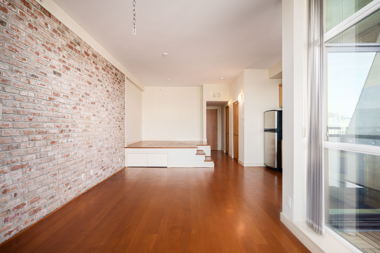 Spacious apartment interior with a brick accent wall, wooden floor, raised platform area, and large windows letting in natural light.