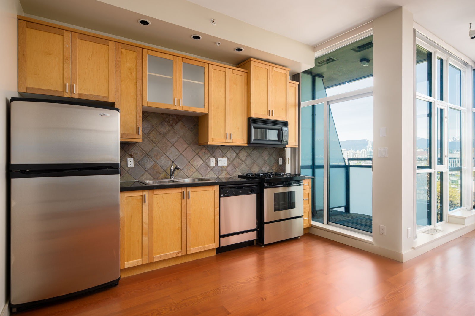 Modern apartment kitchen with light wood cabinets, stainless steel appliances, and tile backsplash. Large windows and a door lead to a balcony with city views.