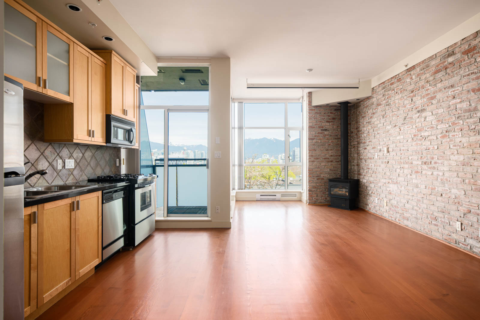Unfurnished apartment with wood flooring, exposed brick wall, modern kitchen, large windows, balcony access, and a view of mountains in the background.