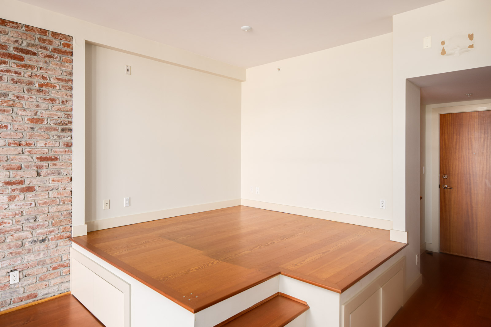 A small, elevated wooden platform with storage drawers in a room featuring a brick accent wall and a wooden door.