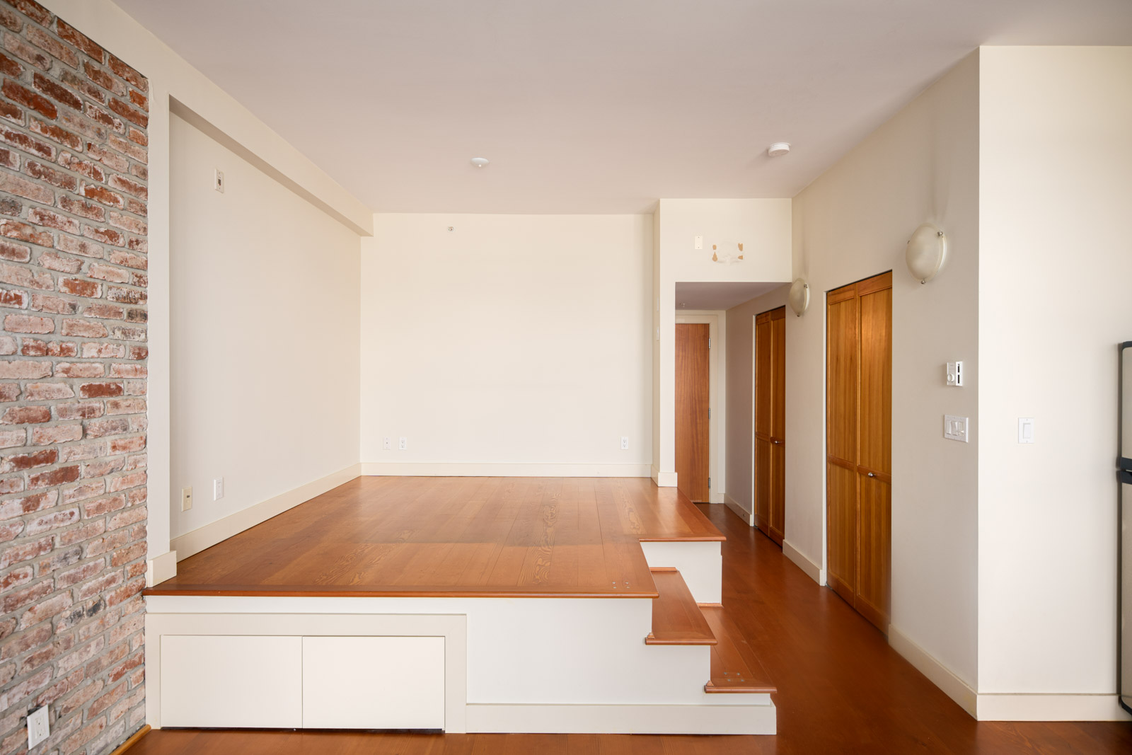 A room with wooden flooring and a raised platform, brick accent wall on the left, and wooden doors along the right hallway.