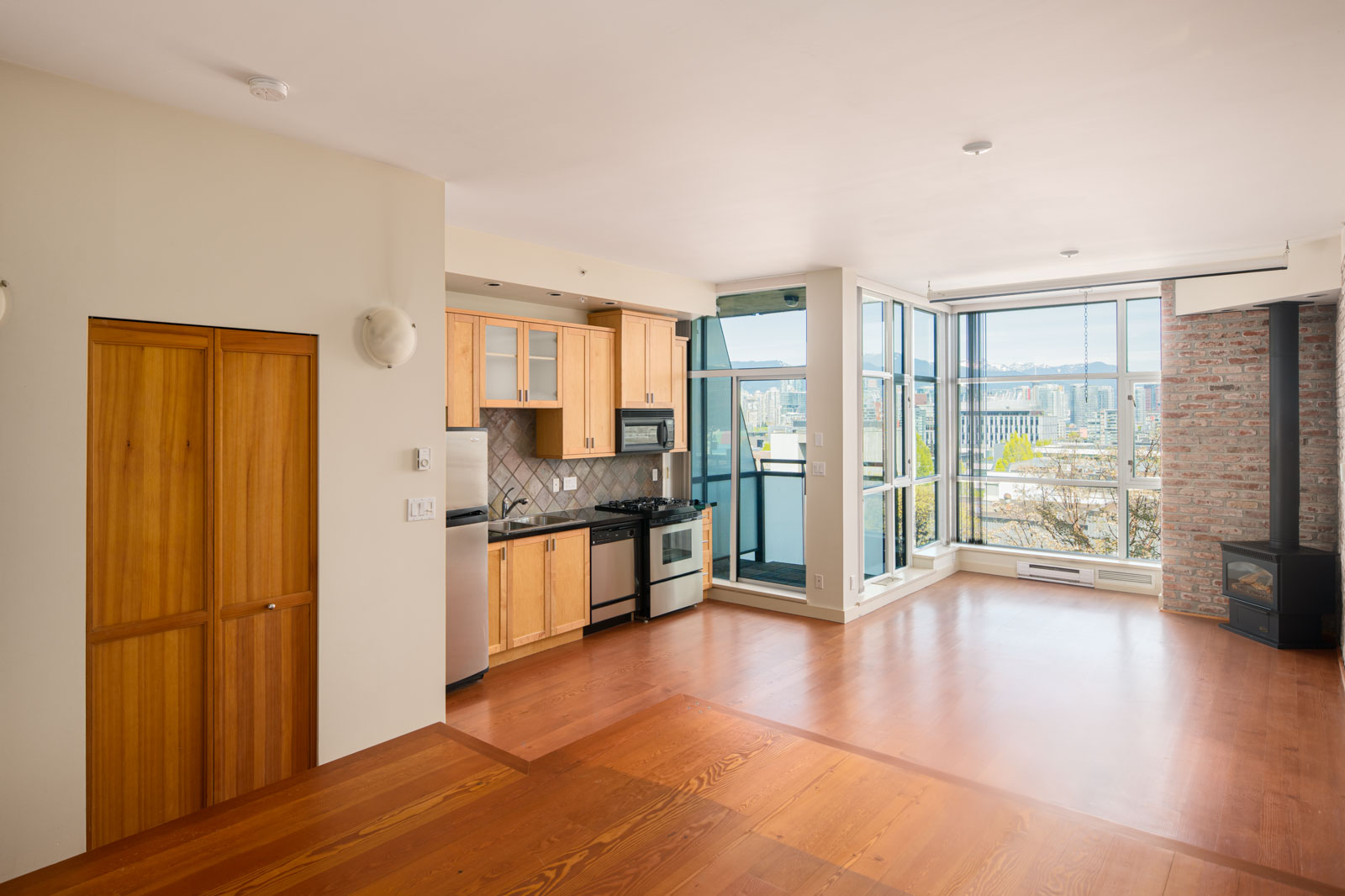 Modern apartment interior with wood floors, open kitchen, large windows, and city view. Features natural light, stainless steel appliances, and exposed brick wall with a fireplace.