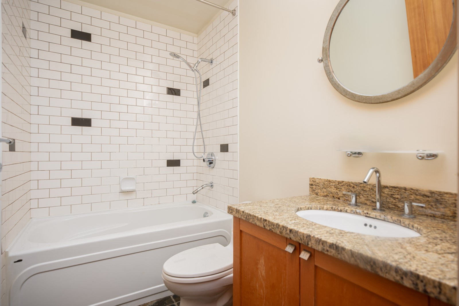 Bathroom with a bathtub and shower, white tile backsplash with black accents, granite countertop sink, and a round mirror above the vanity.