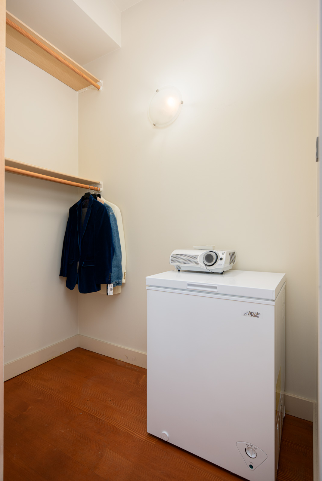 A small closet with a white chest freezer, two jackets hanging on a wooden rod, and a projector on top of the freezer.