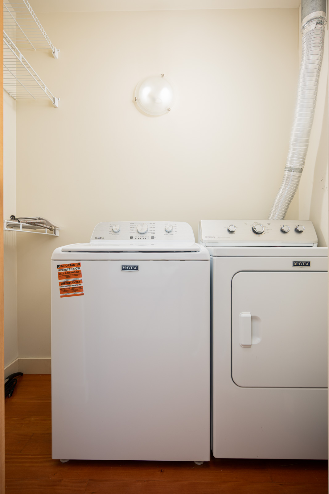 A laundry room with a white Maytag washing machine and dryer side by side, a wall vent hose attached to the dryer, and wire shelving above.