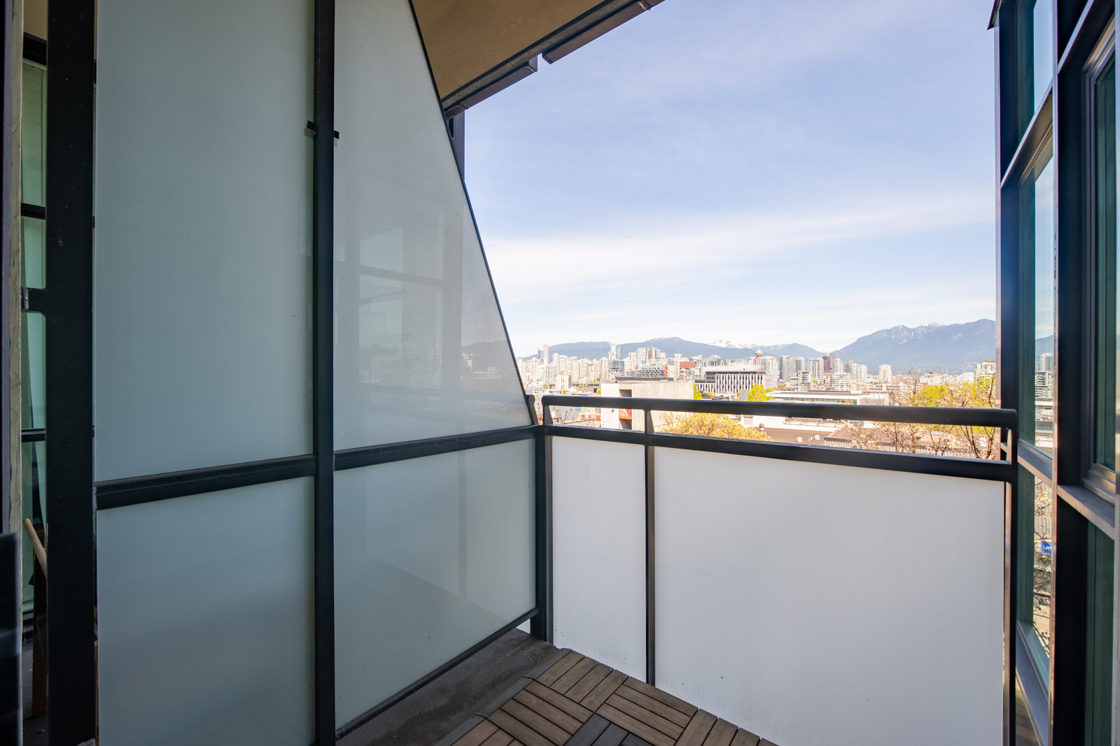 Small modern balcony with frosted glass panels and wood deck tiles, overlooking a cityscape with mountains in the distance under a clear sky.