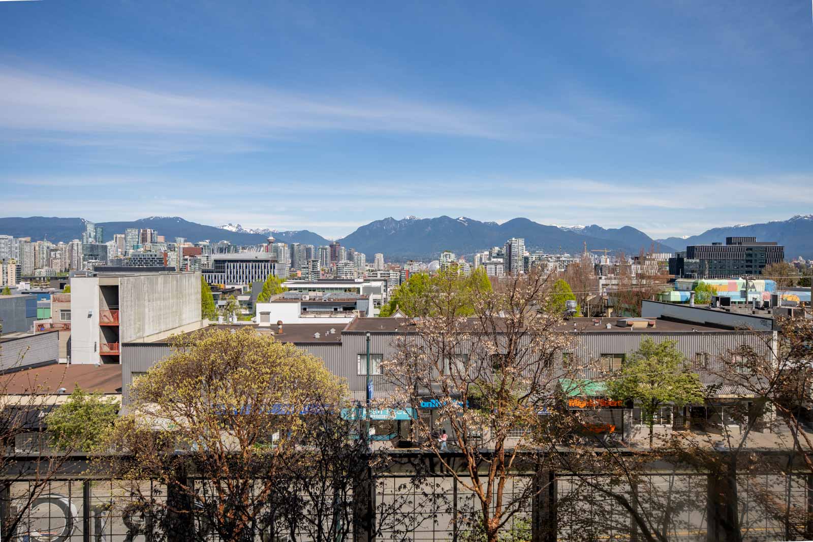 Cityscape with low-rise buildings, trees, and mountains in the background under a blue sky with light clouds.