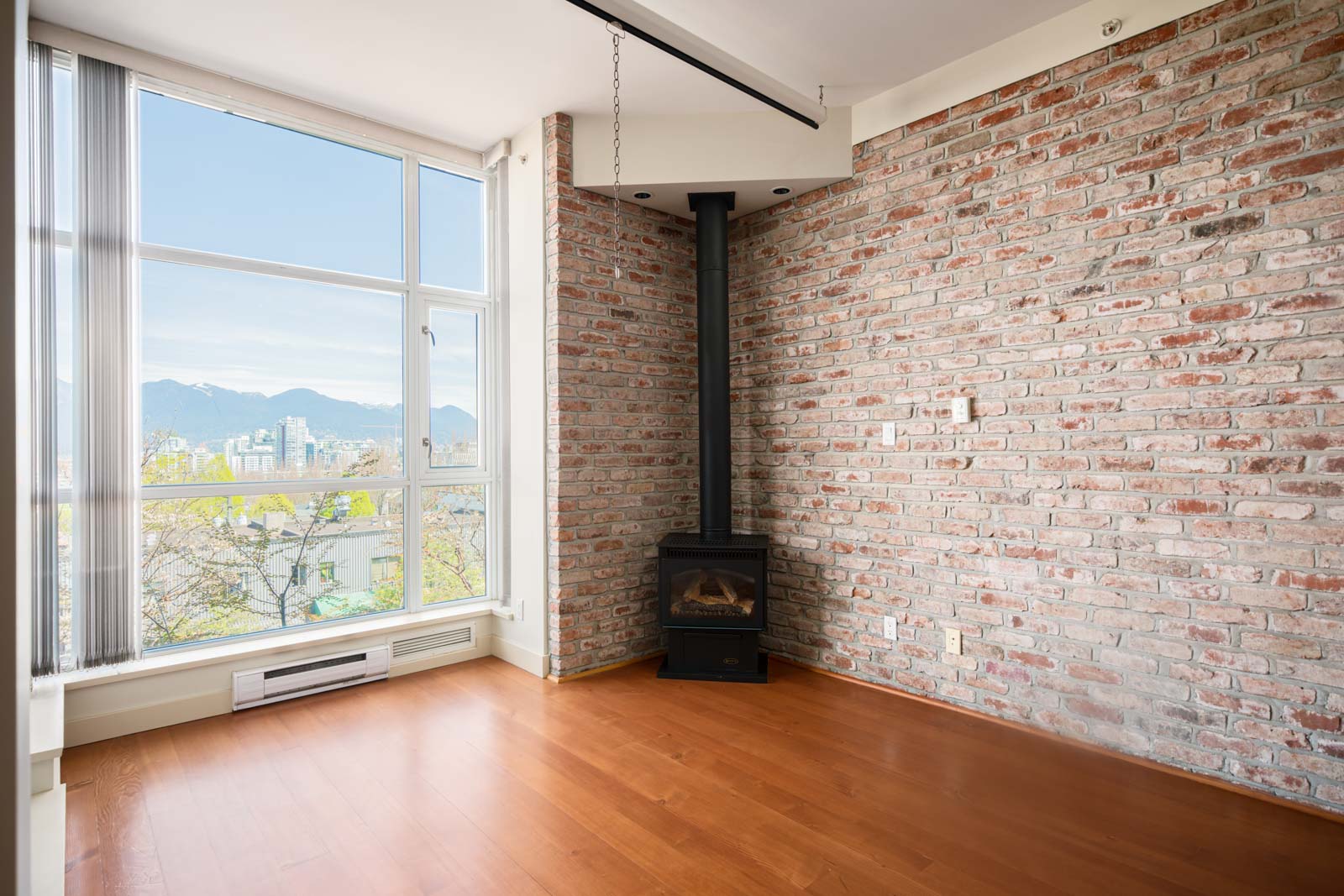 Bright corner room with large windows, exposed brick walls, hardwood floors, and a black wood stove in the corner. Cityscape and mountains visible outside.