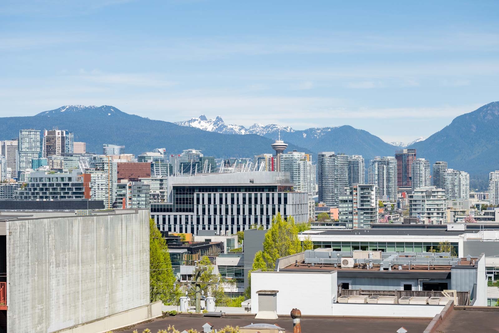Cityscape view of downtown Vancouver with modern buildings in the foreground and snow-capped mountains in the background under a blue sky.