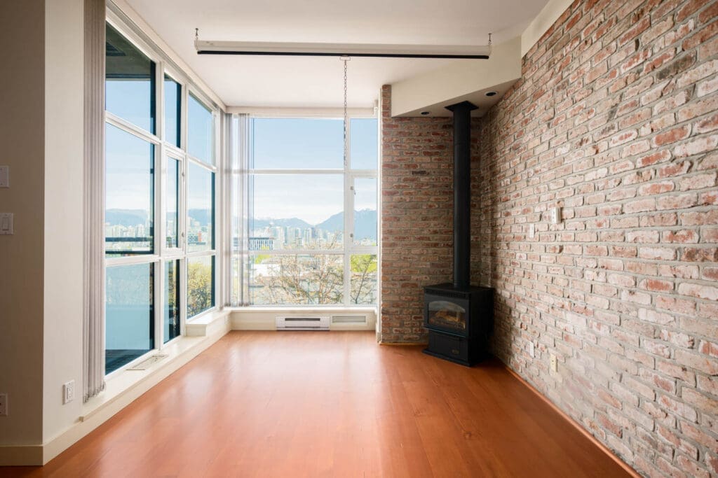 Modern empty room with large floor-to-ceiling windows, exposed brick wall, wood flooring, and a small black wood stove in the corner.